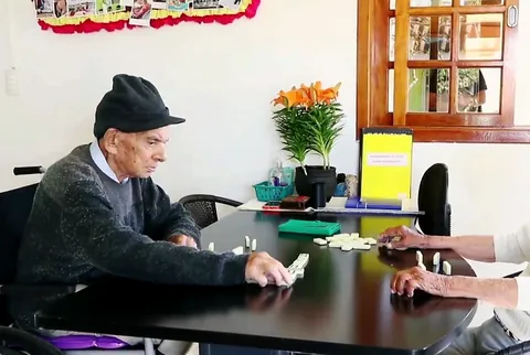 Dois residentes jogam dominó à mesa, com flores e janela ao fundo
