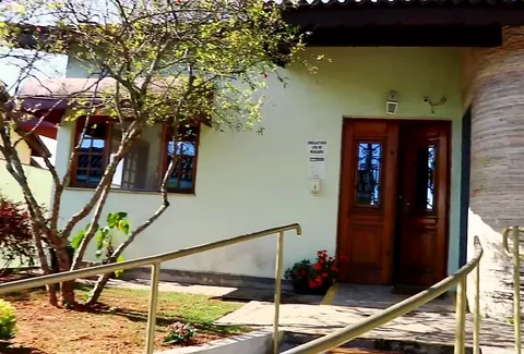 Entrada do Residencial Sol das Olívias, com porta de madeira, parede de pedra, rampa com corrimão e jardim